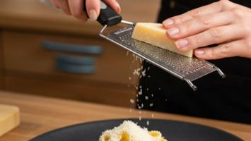 En kok river parmesanost med et rivejern over en tallerken med pasta på et varmt køkkenbord